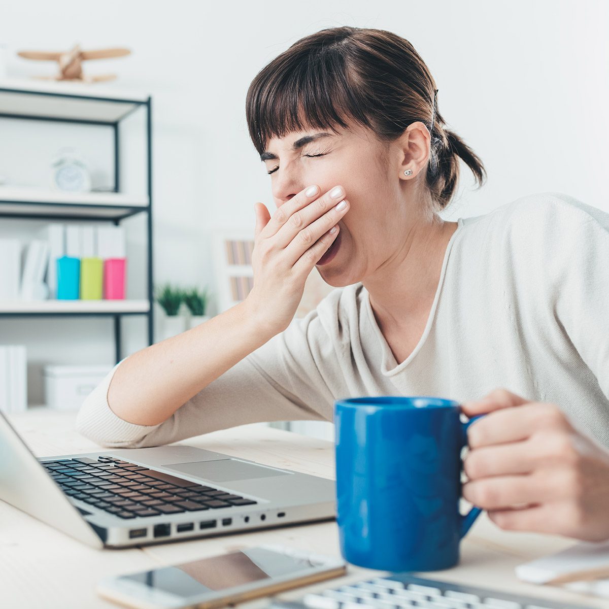 疲倦困倦的女人打着哈欠，在办公桌前工作，端着一杯咖啡，过度工作和睡眠剥夺的概念;伤风ID 367741853