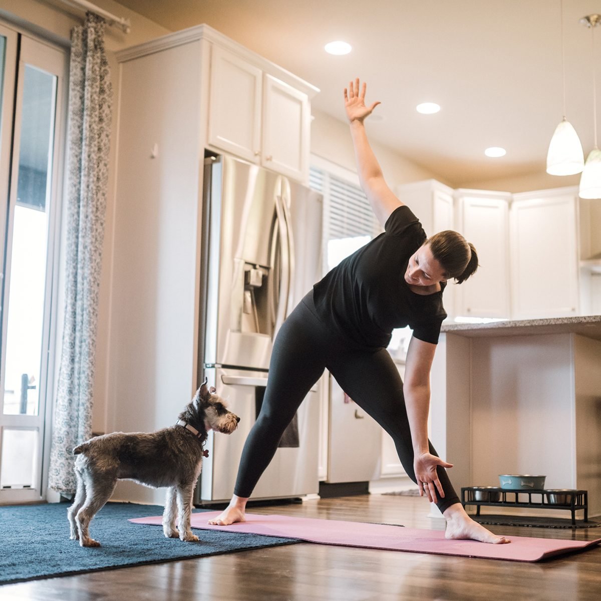 一个成熟的成年妇女在客厅的垫子上做瑜伽和力量训练练习，她的宠物梗狗在她的陪伴下玩耍。